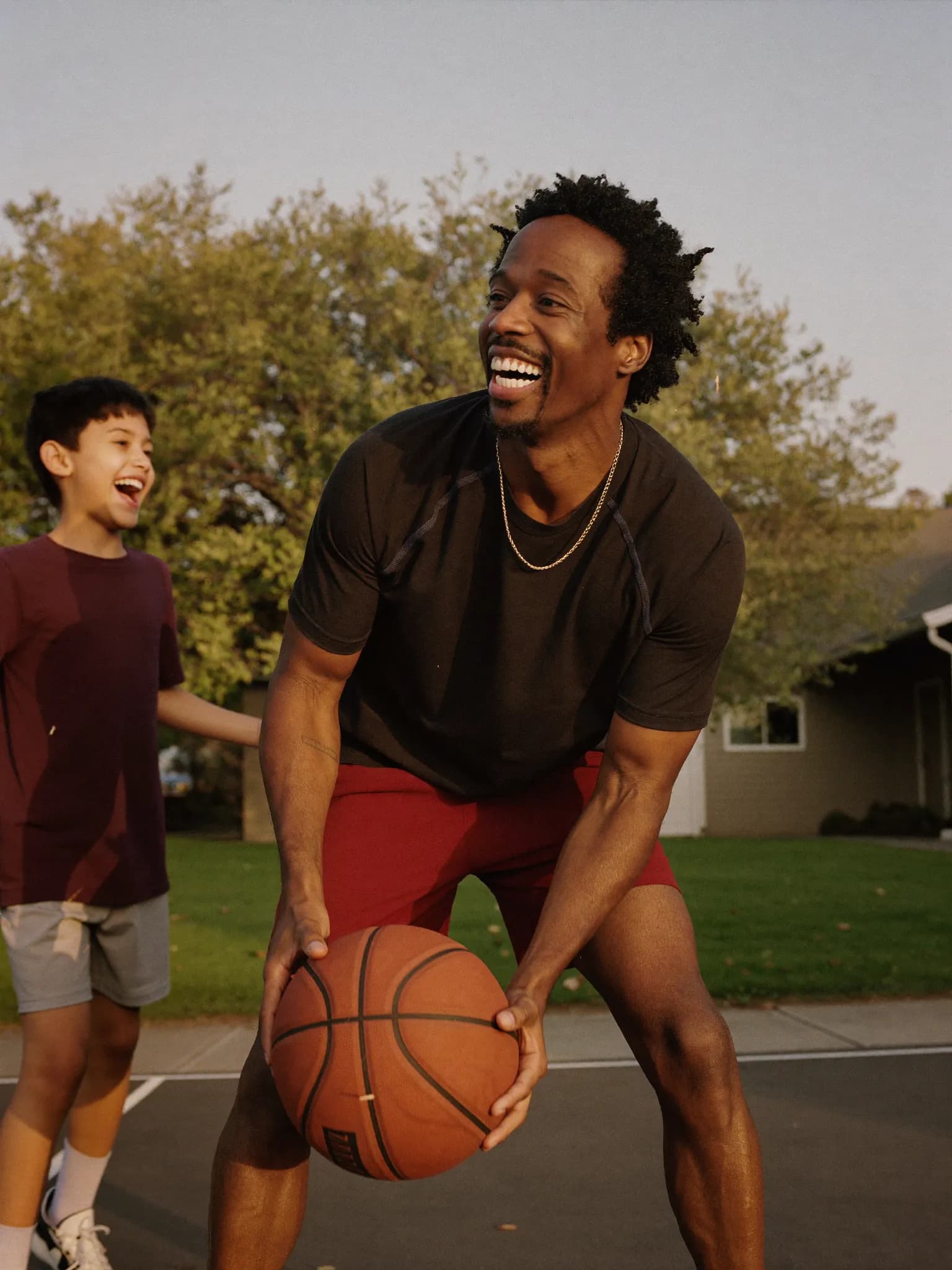A dad in his late 40s laughing while playing driveway basketball with his son at golden hour