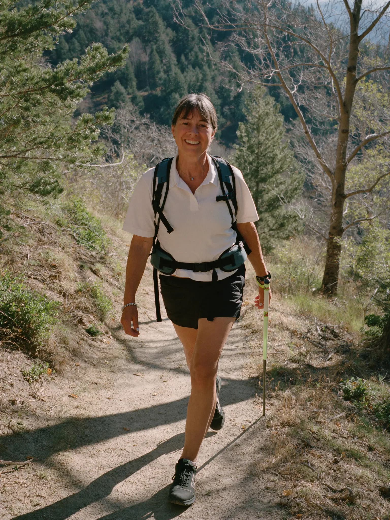 A woman in her early 50s hiking a sunlit trail with an easy, confident stride